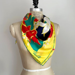 Puede incluir: Un pañuelo de seda amarillo con estampado floral en rojo, verde, blanco y azul. El pañuelo tiene la palabra "Beauty" impresa en verde sobre fondo amarillo.
