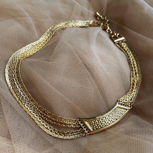 May include: A gold-toned necklace with multiple strands of chain. The necklace features a textured, curved bar at the front and a clasp closure. The jewelry is displayed on a neutral-toned fabric.