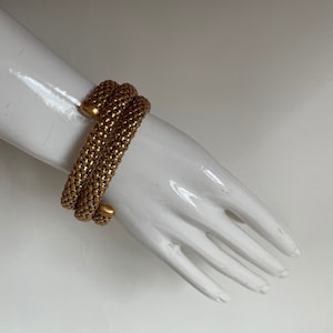 Puede incluir: Un brazalete dorado hecho de múltiples hebras de pequeñas cuentas enlazadas. El brazalete está envuelto alrededor de la muñeca de un maniquí.