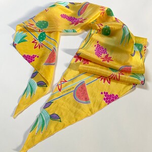 Puede incluir: Un pañuelo de seda amarillo con un estampado colorido de sandía, uvas y cactus. El pañuelo tiene un borde con flecos y el texto "Liz Claiborne" está impreso en el pañuelo.