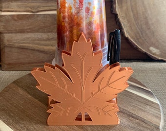 Puede incluir: Un soporte de plástico naranja mate con forma de hoja de arce, que contiene una pila de vasos de plástico transparente decorados con un patrón de hojas de otoño y un bolígrafo negro insertado en el lateral del soporte.