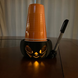 Puede incluir: Una calabaza de pl&aacute;stico negra con una vela encendida en su interior. Un vaso de pl&aacute;stico naranja se coloca encima de la calabaza. Un marcador negro se apoya en la calabaza.