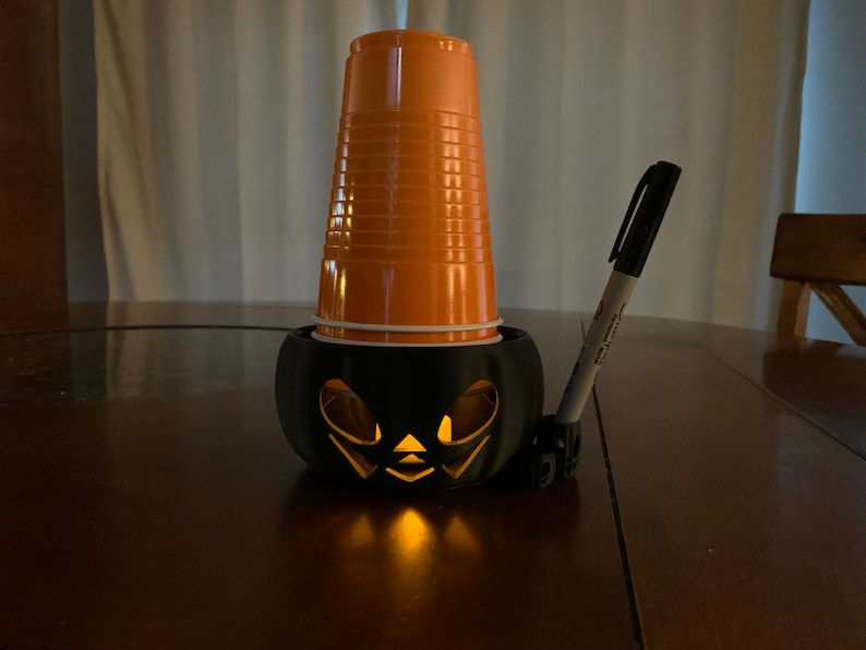 Puede incluir: Una calabaza de pl&aacute;stico negra con una vela encendida en su interior. Un vaso de pl&aacute;stico naranja se coloca encima de la calabaza. Un marcador negro se apoya en la calabaza.