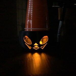 Puede incluir: Una calabaza de Halloween negra con una vela encendida en su interior. La calabaza est&aacute; hecha de un vaso de pl&aacute;stico con una cara tallada.
