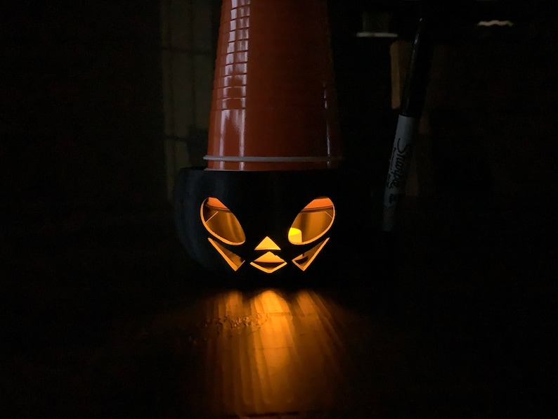 Puede incluir: Una calabaza de Halloween negra con una vela encendida en su interior. La calabaza est&aacute; hecha de un vaso de pl&aacute;stico con una cara tallada.