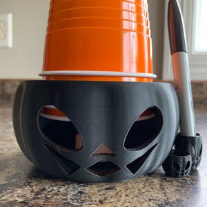 Puede incluir: Un portavasos de pl&aacute;stico negro con forma de calabaza con una cara de calabaza tallada. Un vaso de pl&aacute;stico naranja se encuentra dentro del portavasos. Un marcador negro se apoya en el portavasos.