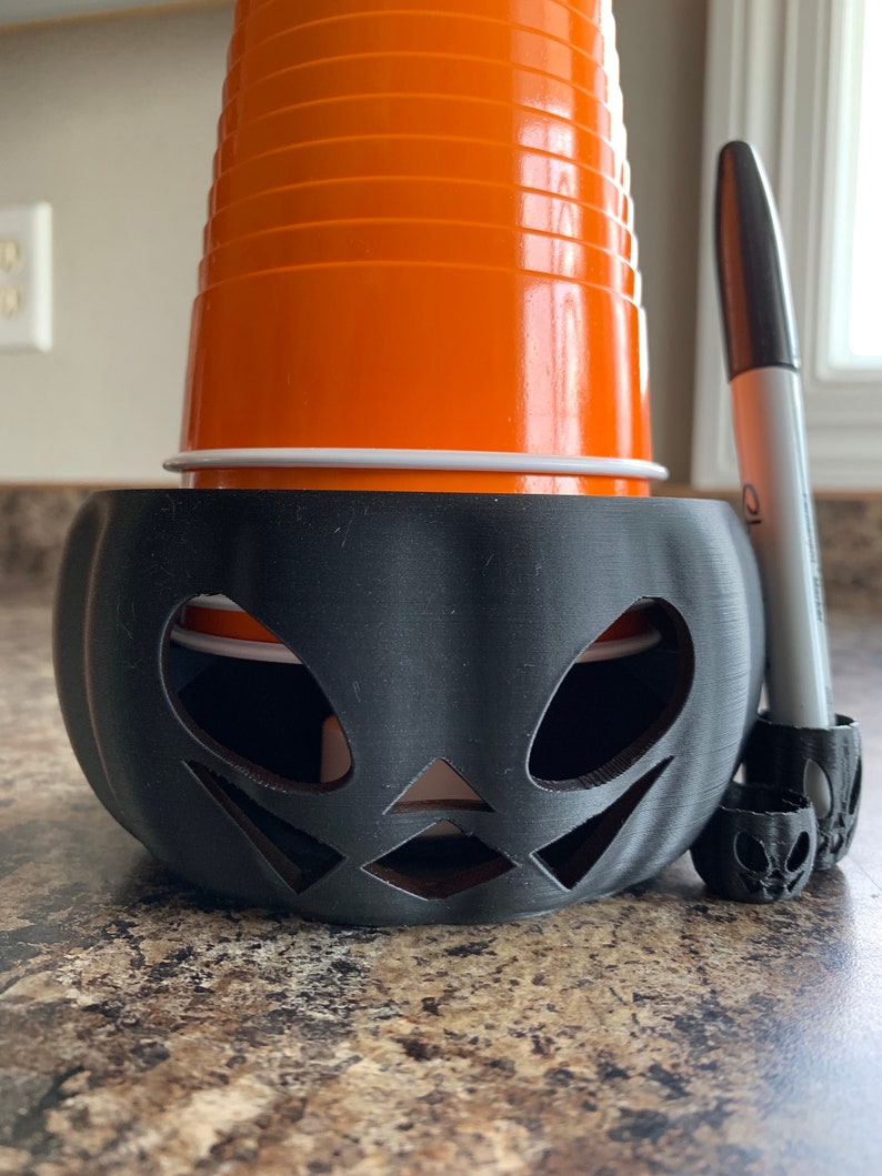 Puede incluir: Un portavasos de pl&aacute;stico negro con forma de calabaza con una cara de calabaza tallada. Un vaso de pl&aacute;stico naranja se encuentra dentro del portavasos. Un marcador negro se apoya en el portavasos.