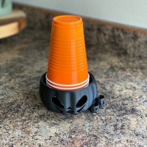 Puede incluir: Un portavasos de pl&aacute;stico negro con forma de calabaza con una cara de calabaza tallada. El portavasos sostiene una pila de vasos de pl&aacute;stico naranja.