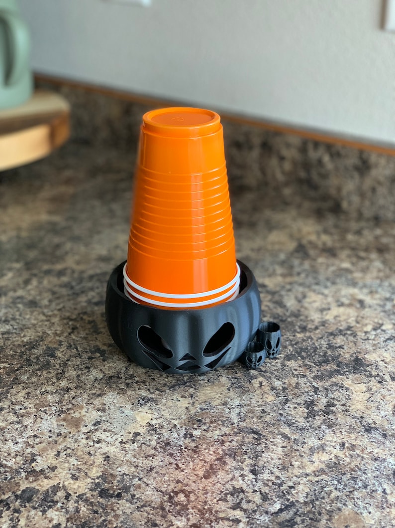 Puede incluir: Un portavasos de pl&aacute;stico negro con forma de calabaza con una cara de calabaza tallada. El portavasos sostiene una pila de vasos de pl&aacute;stico naranja.
