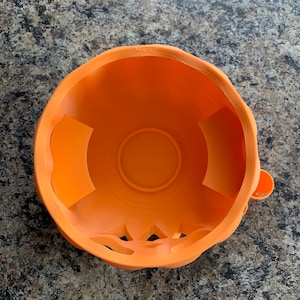 Puede incluir: Taz&oacute;n de pl&aacute;stico naranja en forma de calabaza con un dise&ntilde;o tallado alrededor del borde. El taz&oacute;n tiene un interior liso y redondeado.