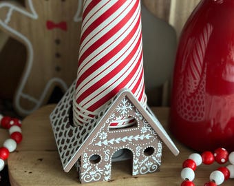 Puede incluir: Un portavasos con forma de casa de jengibre con un vaso de papel a rayas rojas y blancas en su interior. El portavasos está decorado con glaseado blanco y tiene un diseño de copo de nieve en el techo. Hay cuentas rojas y blancas sobre una superficie de madera en primer plano.