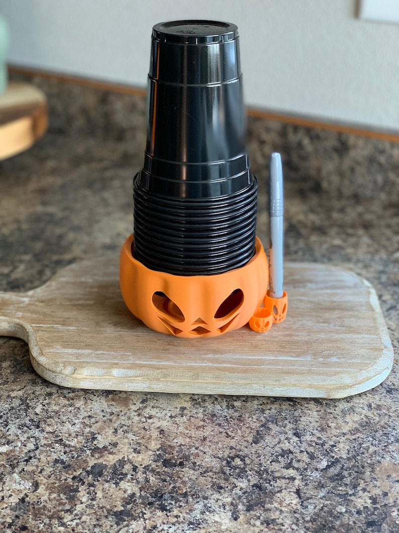 Puede incluir: Un portavasos de pl&aacute;stico negro con forma de calabaza con un bol&iacute;grafo plateado que sobresale de la parte superior. El portavasos est&aacute; sobre una tabla de cortar de madera.