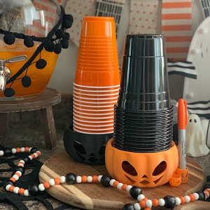 Puede incluir: Portavasos de Halloween de color naranja y negro con forma de calabaza. Los soportes est&aacute;n llenos de pilas de vasos de pl&aacute;stico naranja y negro.