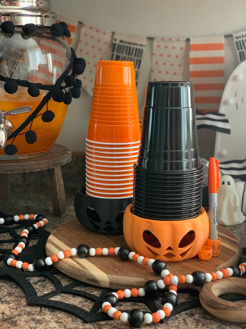 Puede incluir: Portavasos de Halloween de color naranja y negro con forma de calabaza. Los soportes est&aacute;n llenos de pilas de vasos de pl&aacute;stico naranja y negro.