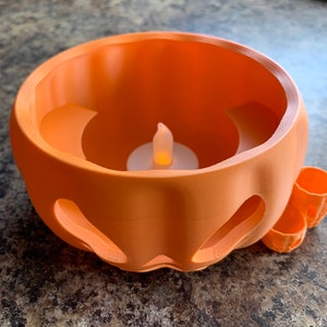 Puede incluir: Un portavelas de pl&aacute;stico naranja en forma de calabaza con una peque&ntilde;a vela LED blanca en su interior. El portavelas tiene una cara tallada y una peque&ntilde;a manija en el lateral.