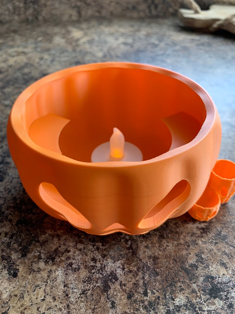 Puede incluir: Un portavelas de pl&aacute;stico naranja en forma de calabaza con una peque&ntilde;a vela LED blanca en su interior. El portavelas tiene una cara tallada y una peque&ntilde;a manija en el lateral.