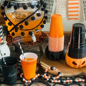 Puede incluir: Una estaci&oacute;n de bebidas para una fiesta de Halloween con un dispensador de vidrio decorado con pompones negros, vasos naranja y negros y una guirnalda de cuentas negras y blancas. El dispensador tiene un cartel que dice "Feliz Halloween".