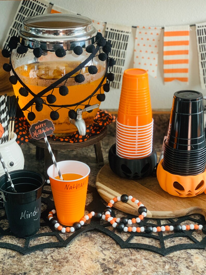 Puede incluir: Una estaci&oacute;n de bebidas para una fiesta de Halloween con un dispensador de vidrio decorado con pompones negros, vasos naranja y negros y una guirnalda de cuentas negras y blancas. El dispensador tiene un cartel que dice "Feliz Halloween".