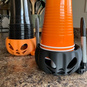 Puede incluir: Dos portavasos de pl&aacute;stico negro con forma de calabaza con vasos de pl&aacute;stico naranja en su interior. Un vaso est&aacute; apilado alto y el otro es un solo vaso.