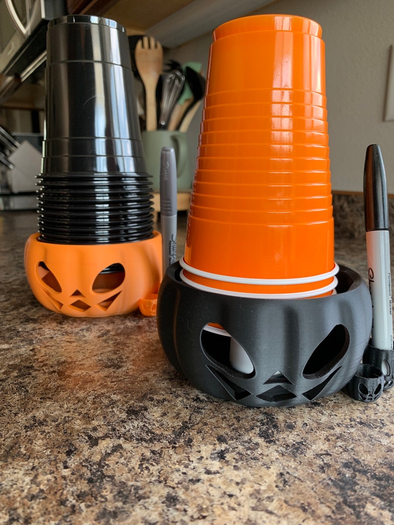 Puede incluir: Dos portavasos de pl&aacute;stico negro con forma de calabaza con vasos de pl&aacute;stico naranja en su interior. Un vaso est&aacute; apilado alto y el otro es un solo vaso.