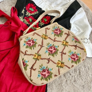 Könnte beinhalten: Eine Vintage-Beige-Handtasche mit einem floralen Nadelspitzendesign mit rosa und blauen Rosen, gelben Blumen und braunen Stielen. Die Tasche hat einen beigen Griff und einen goldenen Verschluss. Die Tasche liegt auf einem roten Kleid, einem schwarzen Oberteil und einer weißen Bluse.