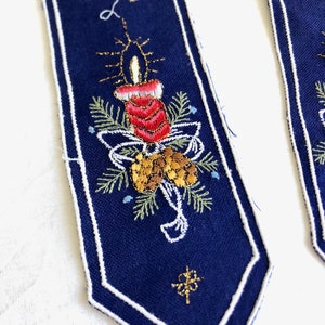 May include: Two blue fabric Christmas decorations with white edging. Each decoration features gold embroidery of a candle, pine branches, pine cones, and the word "Joy".