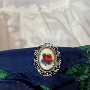 Könnte beinhalten: Silberfarbene Brosche mit ovalem Filigranrahmen. Die Brosche zeigt ein kreuzgesticktes Blumendesign mit roten und blauen Blumen auf weißem Hintergrund. Die Brosche ist auf einem dunkelblauen Band und grünem Stoff platziert.