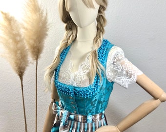 Puede incluir: Un vestido tradicional bávaro azul y blanco con un top de encaje blanco y una falda de cuadros marrones y blancos. El vestido lo lleva un maniquí con el pelo rubio recogido en trenzas.