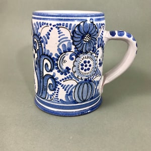 Op de afbeelding: Een witte keramische mok met een blauw en wit bloemenmotief. De mok heeft een handvat en een ronde bodem.