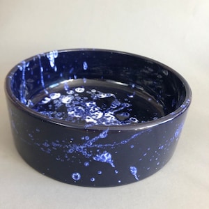 Pode incluir: Uma tigela de cerâmica azul escura com um design de respingos brancos. A tigela tem formato oval e um acabamento liso e brilhante.