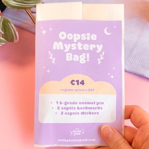 Puede incluir: Una "Oopsie Mystery Bag!" morada con un diseño de nube, luna y estrellas. La bolsa contiene un pin de esmalte de grado B, dos marcadores y tres pegatinas. La bolsa está sujeta por una mano, con un fondo rosa.