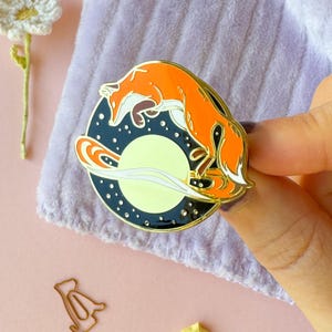 May include: A gold-toned enamel pin featuring a stylized orange fox jumping over a crescent moon with a galaxy design. The pin has a black background with white stars.