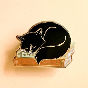May include: A black cat enamel pin sleeping on a stack of books. The pin has gold detailing. The books are in shades of orange and green. A charming accessory.