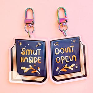 May include: Two acrylic keychains with iridescent clasps. Each keychain is shaped like a book with a navy blue cover and the words "SMUT INSIDE" and "DONT OPEN" in gold lettering. Decorative floral and star accents are included.