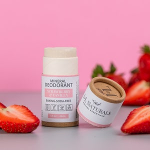 Może przedstawiać: Brązowy kartonowy tubus z białą etykietą, na której widnieje napis "Deodorant mineralny Truskawka & Wanilia Bez sody oczyszczonej" oraz mała zielona ikona liścia. Tubus ma pojemność 1,5 uncji lub 44 ml. Tubus znajduje się obok mniejszego brązowego kartonowego tubusu z różową etykietą, na której widnieje napis "Lil Naturals Ethical Essentials". Oba tubusy znajdują się na szarej powierzchni z pokrojonymi truskawkami.