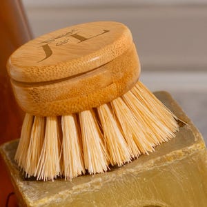 Puede incluir: Un cepillo de bambú para lavar platos con cerdas claras. El mango redondo de madera tiene las iniciales "JL". El cepillo está sobre una pastilla de jabón verde oliva, lo que sugiere productos de limpieza ecológicos.