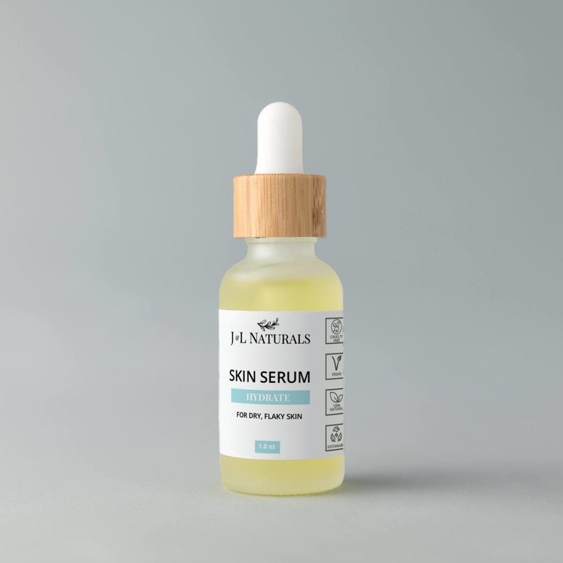 May include: A clear glass bottle with a bamboo lid and a white dropper top. The bottle contains a yellow liquid and has a white label that reads "J+L NATURALS SKIN SERUM HYDRATE FOR DRY, FLAKY SKIN 1.0 oz". The label also has several icons, including a vegan symbol, a cruelty-free symbol, and a natural ingredients symbol.