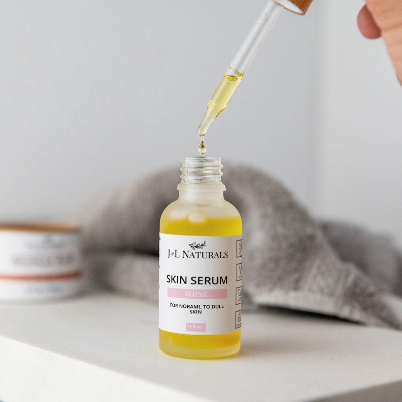 May include: A clear glass bottle containing a yellow liquid. The bottle is labelled "J+L Naturals Skin Serum Shine For Normal to Dull Skin 1.2 oz". A dropper is dispensing the liquid from the bottle.