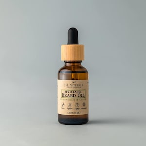 Op de afbeelding: Een bruine glazen fles met een bamboe deksel en een druppelaar. De fles bevat baardolie met geuren van citroengras en kruidnagel. Het etiket vermeldt "J-L Naturals Ethical Essentials Hydrate Beard Oil Lemongrass & Clove" en bevat pictogrammen voor veganistisch, paraben-vrij, dierproefvrij en duurzaam. De fles is 30 ml.