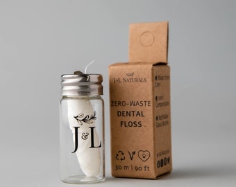 May include: A glass dental floss container with a silver lid and white floss. The container has a black floral design and the letters "J & L". A brown box next to it reads "ZERO-WASTE DENTAL FLOSS", with measurements of 30 ml / 90 ft.