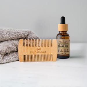 May include: A wooden beard comb and a bottle of beard oil are displayed on a white surface. The comb is light brown with the text "J+L NATURALS". The beard oil bottle is brown with a black dropper and a label that reads "HYDRATE BEARD OIL". A gray towel is in the background.