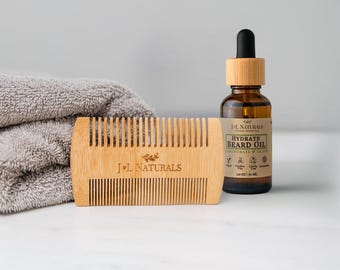 May include: A wooden beard comb and a bottle of beard oil are displayed on a white surface. The comb is light brown with the text "J+L NATURALS". The beard oil bottle is brown with a black dropper and a label that reads "HYDRATE BEARD OIL". A gray towel is in the background.