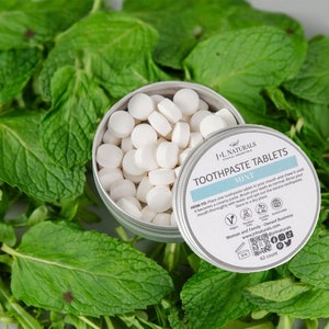 Op de afbeelding: Een zilveren blikje gevuld met witte tandpasta tabletten, met het label "J-L Naturals Toothpaste Tablets Mint". Het blikje staat op een bed van groene muntbladeren.