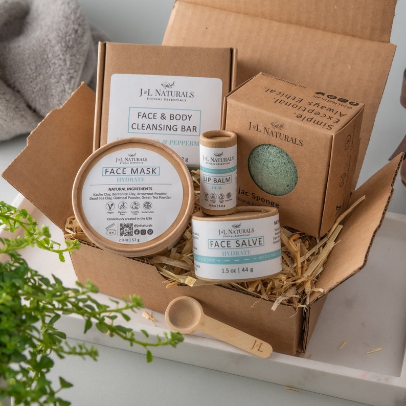 K&ouml;nnte beinhalten: Eine Geschenkbox gef&uuml;llt mit nat&uuml;rlichen Hautpflegeprodukten, darunter eine Gesichtsmaske, ein Lippenbalsam, eine Gesichtscreme und ein Konjak-Schwamm. Die Produkte sind in braunen Pappkartons und -r&ouml;hrchen mit wei&szlig;em Text und einem hellblauen Akzent verpackt. Die Box ist mit zerkleinertem Papier gef&uuml;llt.
