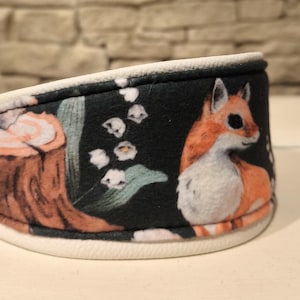 Könnte beinhalten: Ein schwarzes Stirnband mit einer Waldszene, die ein Eichhörnchen, einen Baumstumpf und Maiglöckchenblumen zeigt. Das Stirnband hat eine weiße Innenseite und eine schwarze Außenseite mit dem illustrierten Design.