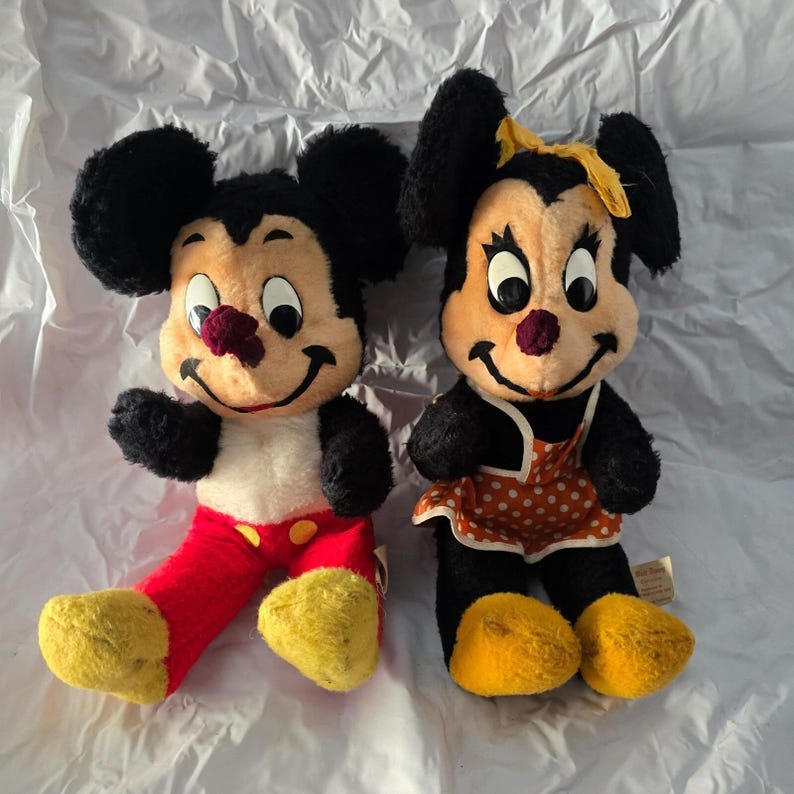 May include: Two vintage Mickey and Minnie Mouse plush dolls. Mickey wears red pants with yellow buttons and shoes. Minnie wears a polka-dot dress and yellow bow. Both have black ears, smiling faces, and are sitting.