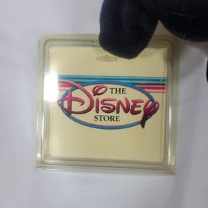 May include: A clear plastic tag with the Disney Store logo in pink, red, and blue. The tag is attached to a black plush toy. The logo features the word "Disney" in a stylized script font.