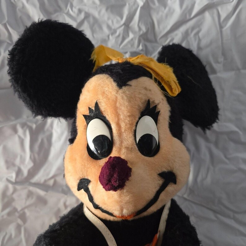 May include: Vintage plush toy featuring a mouse-like character with large black ears, a peach-colored face, and a purple nose. The character has large white eyes with black pupils, black eyelashes, and a yellow bow. The toy is black and peach-colored.