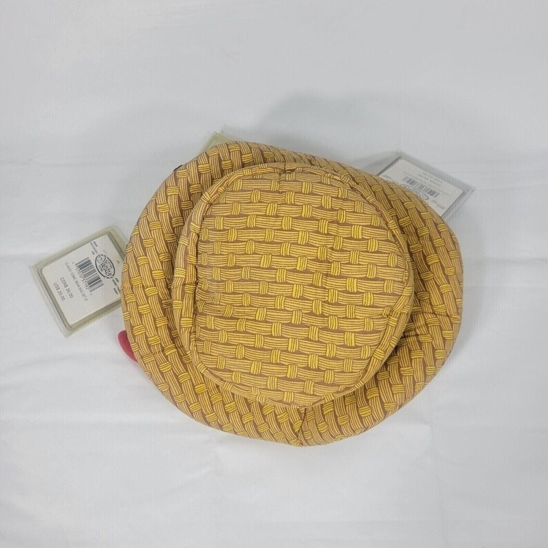 May include: A woven-patterned hat in shades of yellow and brown, with a rounded crown and brim. The hat has a basket-weave design. Two tags are attached to the hat. The hat is sitting on a white surface.