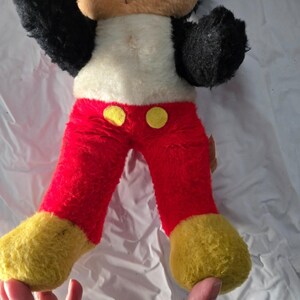 May include: Vintage Mickey Mouse plush toy. The plush features a white torso, red pants with yellow buttons, black arms, and yellow feet. The toy's arms are raised, and it appears to be in good condition for its age.